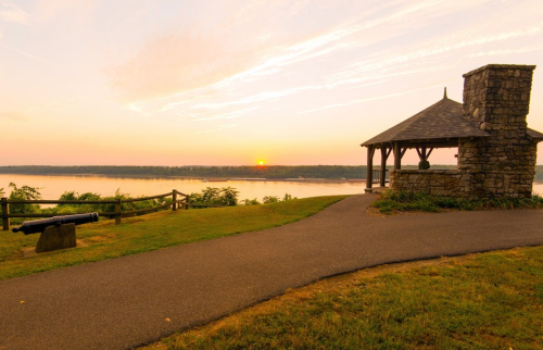 A serene riverside scene at sunset, featuring a gazebo, a cannon, and a winding path along the water.