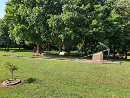 A modern sculpture in a grassy park, surrounded by trees, with a small tree and informational sign nearby.