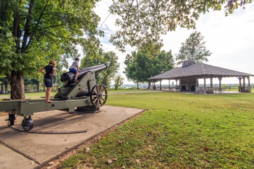 Two children play on a historic cannon in a park, with a gazebo and trees in the background.