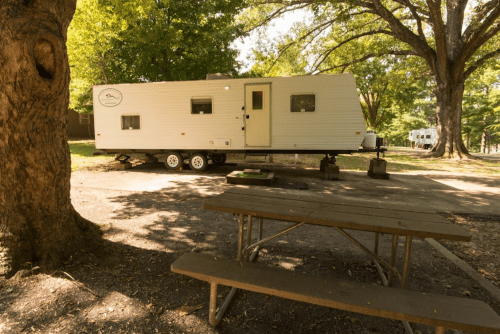 A white trailer parked in a shaded area with a picnic table nearby, surrounded by trees.