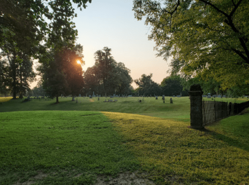 A serene cemetery at sunset, with green grass, trees, and gravestones visible in the distance.