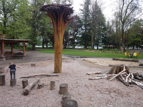 A playground with wooden structures, logs, and a tall tree-like feature, surrounded by greenery and a path.