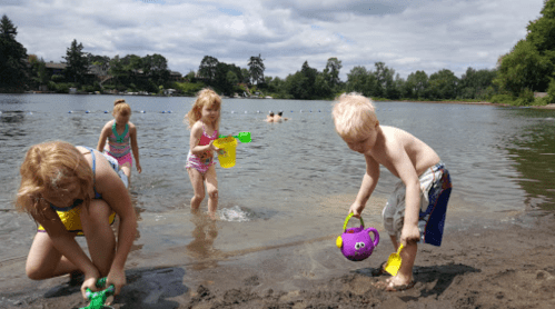 Children playing at a lake, using colorful buckets and toys in the water on a sunny day.