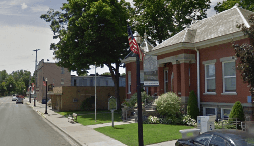 A street view of a red brick building with trees, benches, and American flags along the sidewalk.