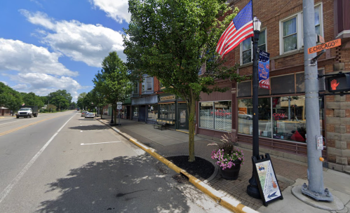 A quiet street scene featuring shops, trees, and an American flag on a sunny day with clouds in the sky.