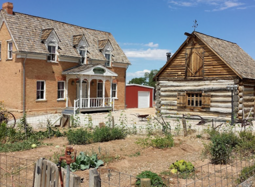 A rustic scene featuring a brick house and a log cabin, surrounded by a garden under a clear blue sky.