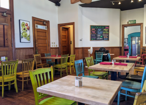 Colorful chairs surround wooden tables in a cozy restaurant with a chalkboard menu on the wall.