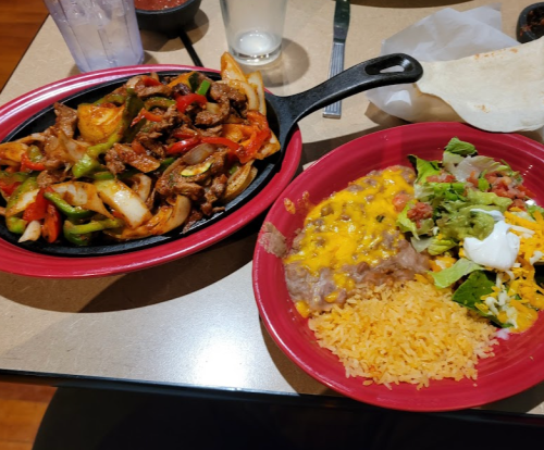 A sizzling platter of fajitas with peppers and onions, alongside rice, refried beans, and a salad with cheese and sour cream.