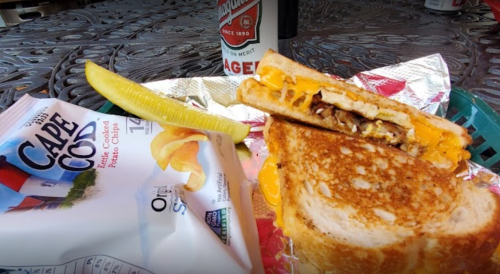 A grilled cheese sandwich with melted cheese, a pickle, and a bag of Cape Cod chips on a patterned table.