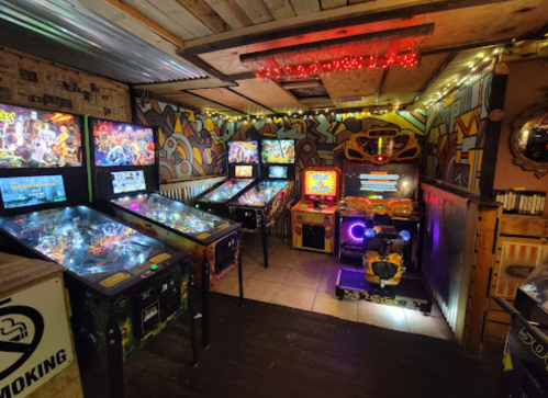 A cozy arcade room featuring several pinball machines and a racing game, with colorful walls and ambient lighting.