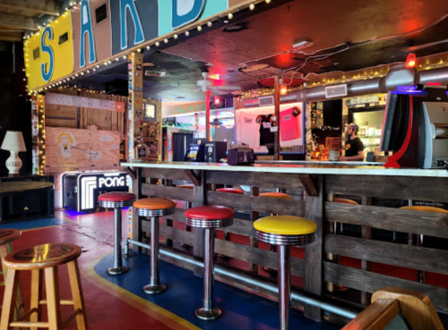 A vibrant bar area with colorful stools, neon lights, and arcade games in a lively setting.