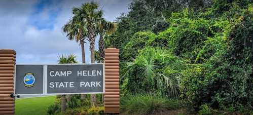 Sign for Camp Helen State Park surrounded by lush greenery and palm trees under a cloudy sky.