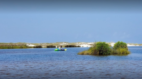 A person kayaking in a calm waterway surrounded by lush greenery and sandy shores under a clear blue sky.