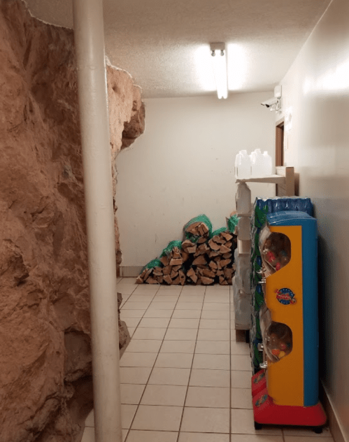 A narrow hallway with a rock wall, stacked firewood, and a colorful vending machine on the right.