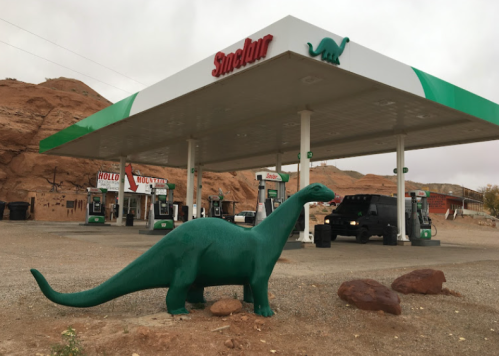 A green dinosaur statue stands in front of a Sinclair gas station amidst rocky terrain.