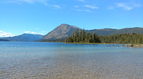 A serene lake surrounded by mountains and lush trees under a clear blue sky.