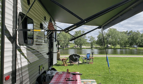 A parked RV by a lake, with a picnic setup and chairs under an awning on a grassy area.