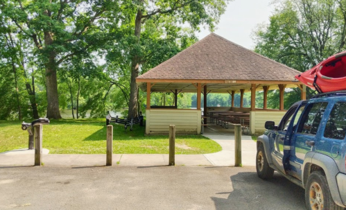 A gazebo by a river, surrounded by trees, with a parked vehicle and kayak on top.