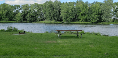 A peaceful riverside scene featuring a picnic table on green grass, surrounded by trees and calm water.