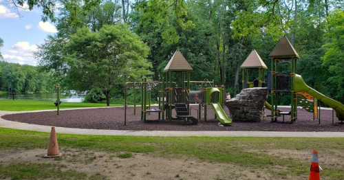 A playground with slides and climbing structures, surrounded by trees and a grassy area near a calm lake.