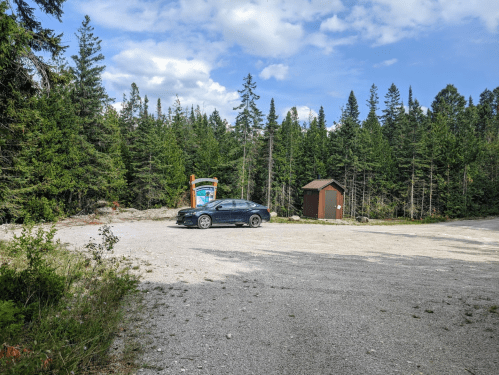 A black car parked in a gravel area surrounded by dense green trees and a small wooden structure.