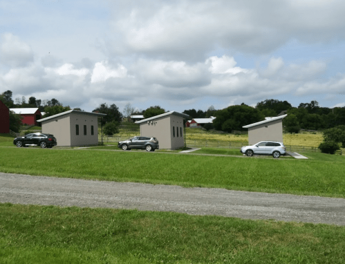 Three small modern buildings with cars parked in front, set in a grassy area with trees and a cloudy sky in the background.