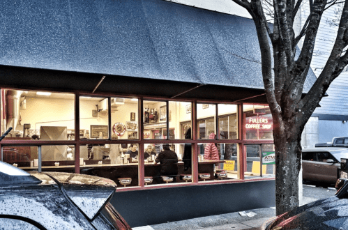 A cozy coffee shop with large windows, showing patrons inside and a tree outside, parked cars in the foreground.