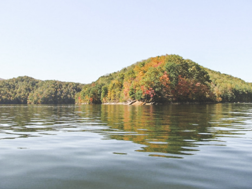 A serene lake scene with a colorful, tree-covered island reflecting on the calm water under a clear sky.