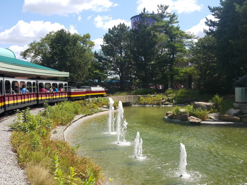 A colorful train passes by a pond with fountains, surrounded by greenery and trees under a blue sky.