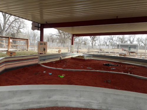A spacious outdoor area with red sand, surrounded by a covered structure and fenced enclosures for animals.