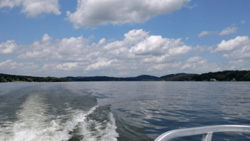 A serene lake view with gentle waves, under a blue sky filled with fluffy clouds and distant hills.
