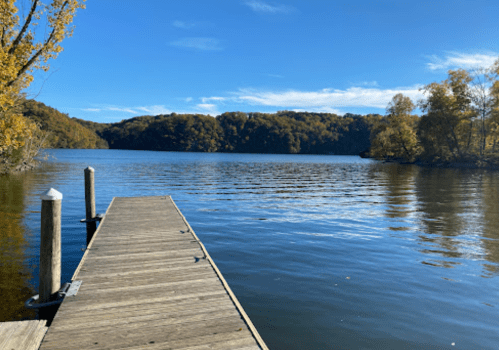 A wooden dock extends into a calm lake surrounded by autumn-colored trees under a clear blue sky.