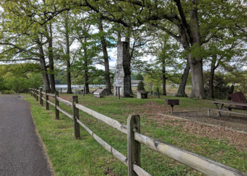 A grassy area with a stone chimney, picnic tables, and trees along a path near a river.