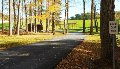 A paved path leads through a wooded area with autumn leaves, picnic shelters, and a sign for a group camp area.