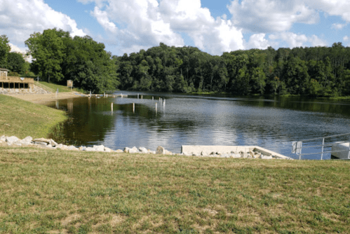 A serene lake surrounded by greenery, with a grassy shore and wooden structures in the background under a cloudy sky.