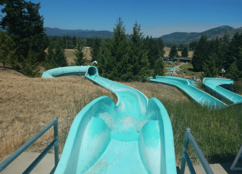 Two turquoise water slides winding down a grassy hill, surrounded by trees and mountains in the background.
