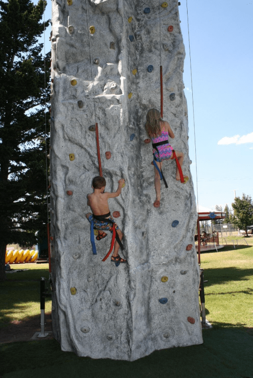 Two children climbing a rock wall at a park on a sunny day, wearing harnesses and colorful climbing gear.