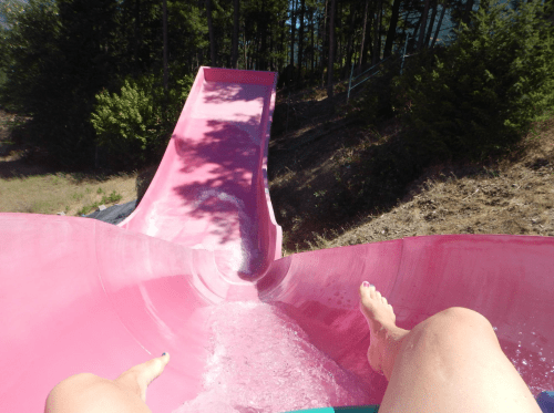 A view from the top of a pink water slide, with feet visible and trees in the background.
