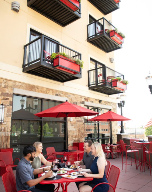 A group of four people dining outdoors at a restaurant with red umbrellas and balcony planters in the background.