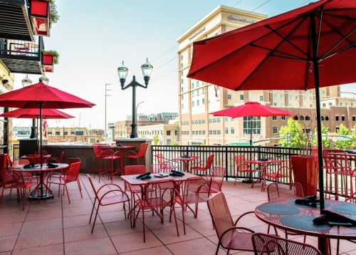 Outdoor patio with red tables and umbrellas, overlooking a cityscape with buildings and streetlights.
