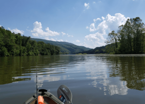 A serene view of a river surrounded by lush green hills and blue skies, seen from a kayak.