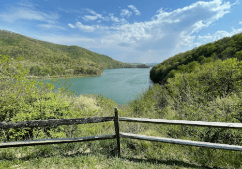 Scenic view of a river winding through green hills under a blue sky with fluffy clouds, framed by a wooden fence.
