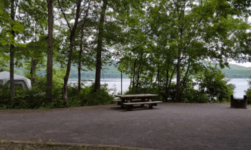 A serene lakeside campsite surrounded by trees, featuring a picnic table and a fire pit.