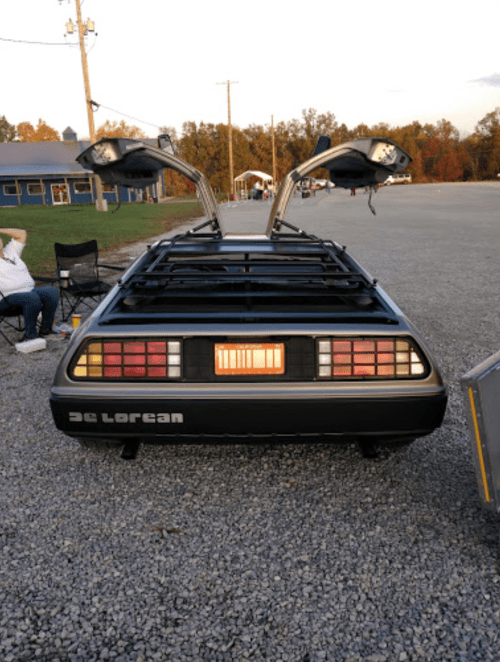 A DeLorean car with its gullwing doors open, parked on gravel with a rural background and a person sitting nearby.