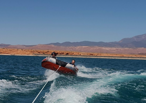 Two people enjoy tubing on a lake, being pulled by a boat, with mountains in the background under a clear blue sky.