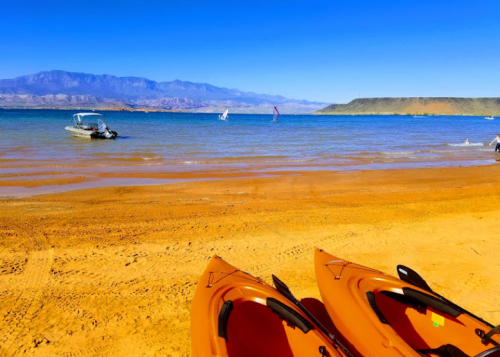 Two kayaks on a sandy beach with a calm lake, boats in the distance, and mountains under a clear blue sky.