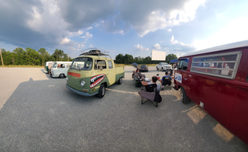 A green vintage van with a shark mouth design parked near people sitting in chairs, surrounded by other vehicles.