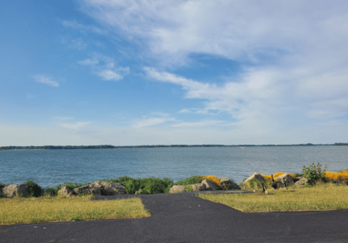 A serene view of a calm lake with a clear blue sky and grassy shoreline, framed by rocks and distant trees.