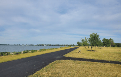 A peaceful lakeside view with a paved path, grassy areas, and trees under a partly cloudy sky.