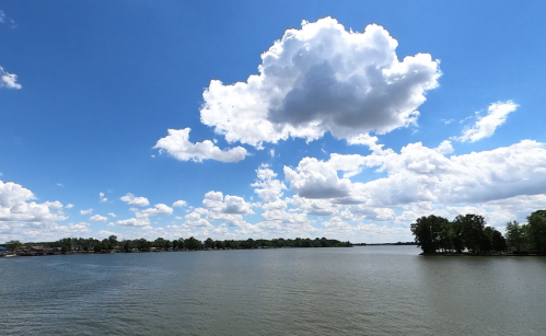 A serene lake under a bright blue sky filled with fluffy white clouds, surrounded by trees along the shoreline.